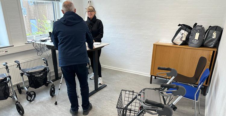 En mand med stok står over for en kvindelig medarbejder ved et hæve-sænkebord i et lyst lokale. Rundt om står to rollatorer, en rollatorkurv og hjælpemiddeltasker.